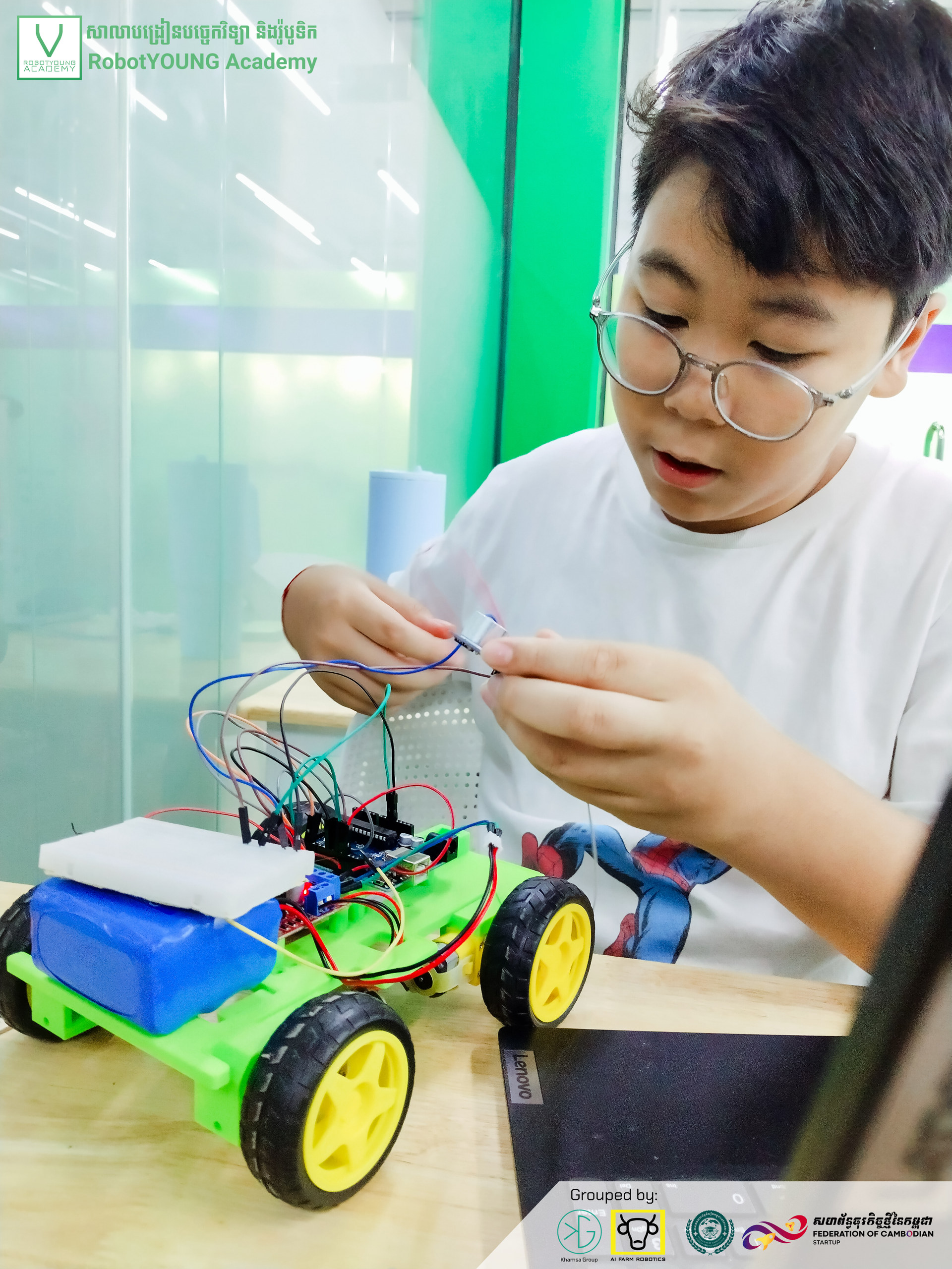 A student wearing glasses connecting wires on a green robot with yellow wheels