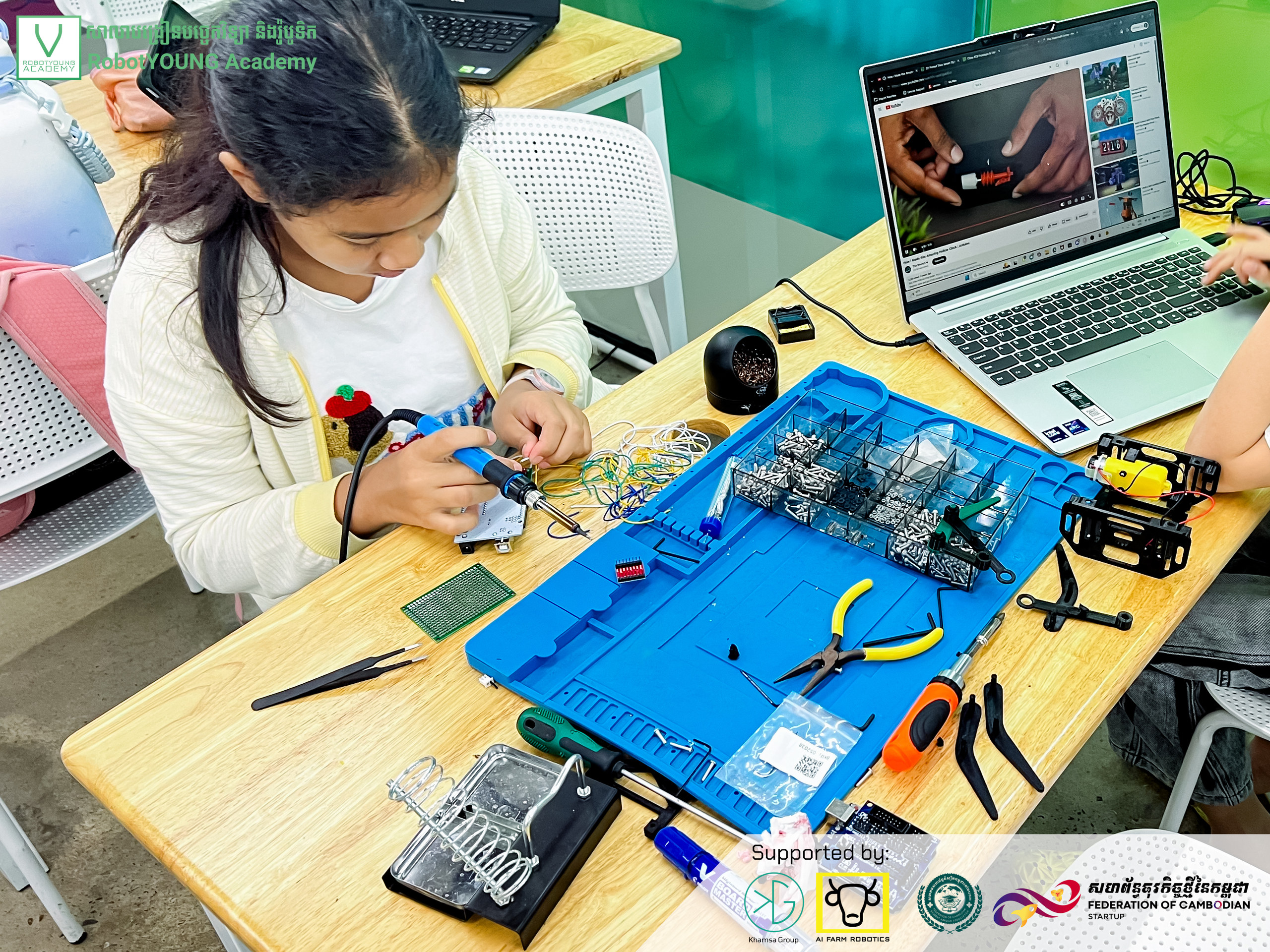 A learner working on a circuit board on a blue electronics mat beside a laptop