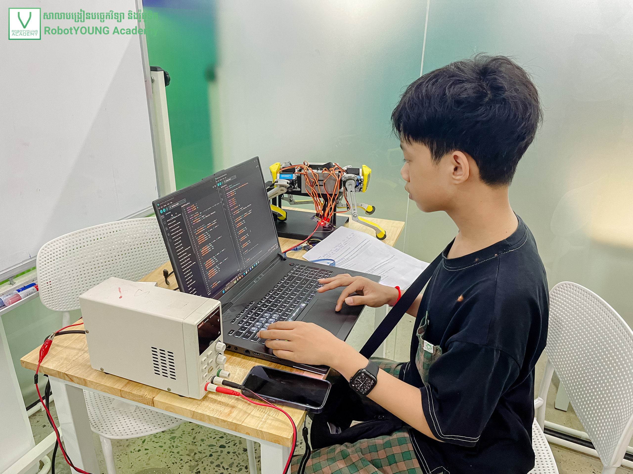 A student typing code on a laptop beside a wheeled robot and white enclosure