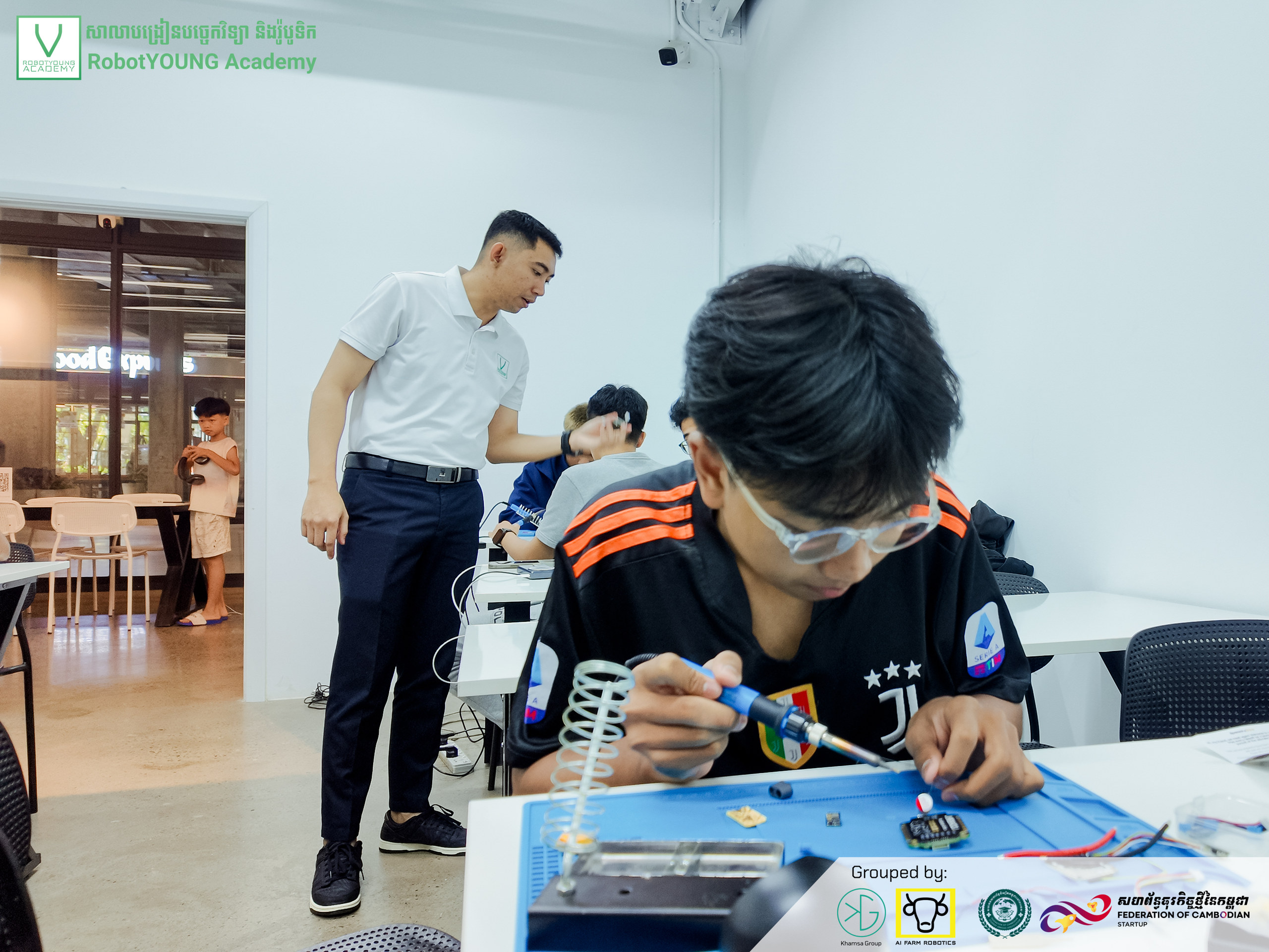 A learner concentrating on a circuit board on a blue work mat while an instructor stands nearby