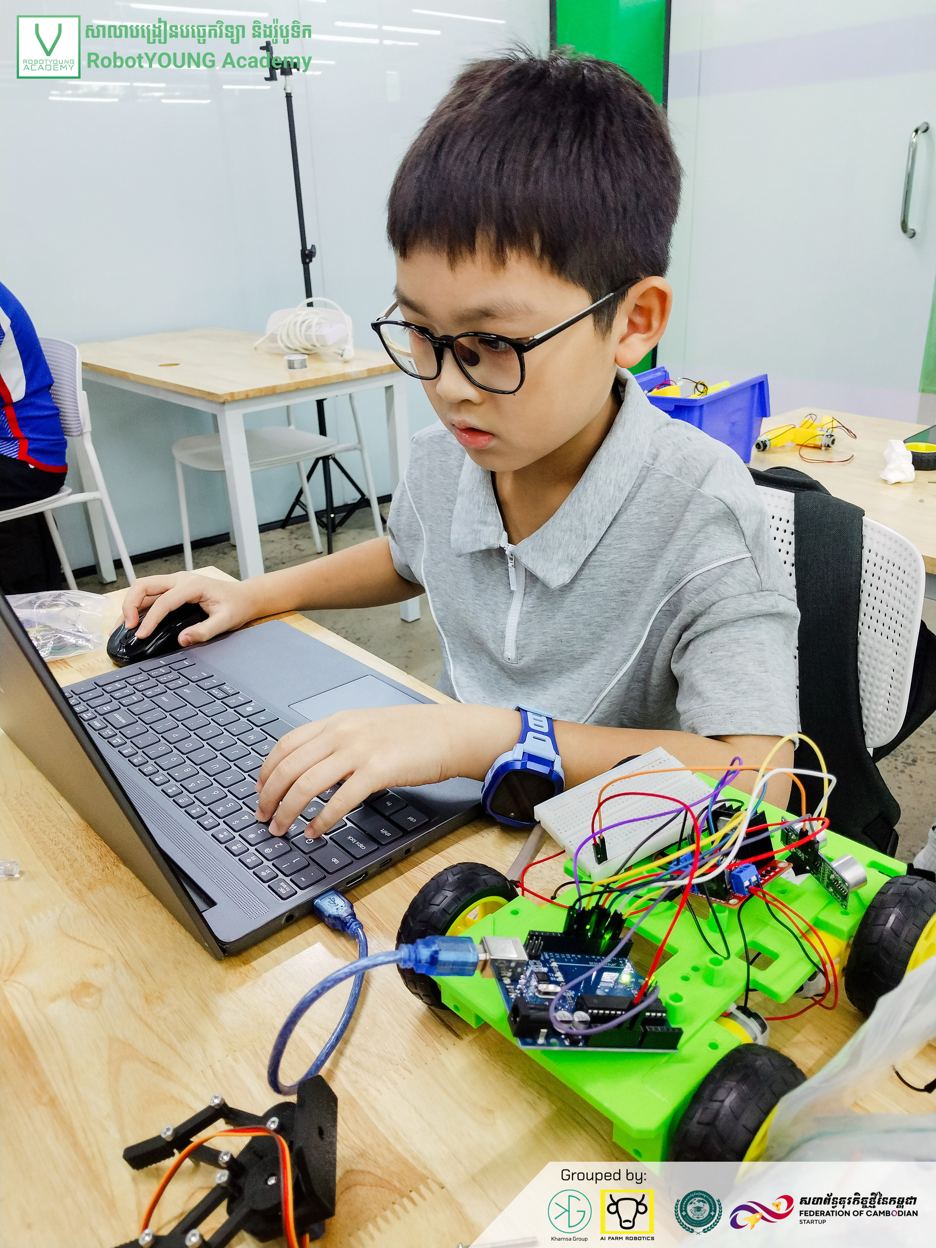 A student wearing glasses typing on a laptop beside a green wheeled robot